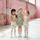 Three young girls in matching outfits holding tennis rackets in a pink industrial setting.