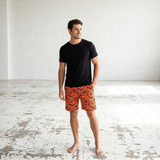 Man wearing black t-shirt and orange patterned shorts standing in a large, empty room with white walls and a concrete floor.