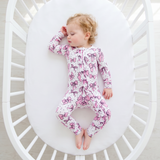 A baby lying on their back inside a white oval crib, wearing a long-sleeve footless pajama romper with a pink and purple unicorn pattern on a white background.