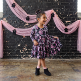 Young girl in a floral dress standing in front of a black brick wall with pink drapes and Halloween decorations.