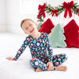 A young boy wearing a festive Christmas pajama set with a colorful holiday print, sitting on a white bed in a bright, white-walled bedroom decorated with a green garland and red bows.