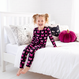 A young girl with blonde pigtails sits on a white bed wearing a black long-sleeve pajama set with a pink bow pattern. The room is brightly lit with white walls and a white wooden bed frame, decorated with spiderweb pillows, a black cat pillow, and a magenta pumpkin pillow.