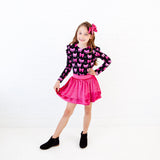 A young girl modeling a long-sleeve black top with a pink butterfly pattern and a bright pink pleated skirt, paired with a pink bow in her hair, standing against a white brick wall.
