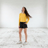 Woman in a yellow sweater and black shorts standing in a white room.