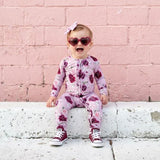 Child wearing a pink floral outfit sitting on a ledge against a pink brick wall.