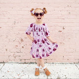 Young girl in a floral dress standing against a light pink brick wall.