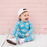 Child wearing a blue onesie with pink patterns, sitting on a white surface against a pink brick wall.