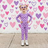 Child in purple polka dot outfit standing in front of a heart-patterned wall.