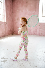Young girl in a floral outfit holding a green tennis racket in a pink room.