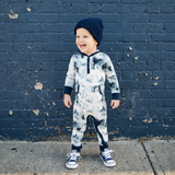 Child wearing a tie-dye onesie and navy hat standing against a blue brick wall.