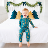 A young child wearing a teal and mustard yellow Christmas tree print zip-up pajama set sits on a white bed with holiday green garland and tree pillows in the background.