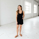 Woman in a black romper standing in a bright, white room.