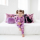 Child in pink Halloween-themed pajamas sitting on a white couch with decorative pillows and a pumpkin.