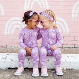 Two children in matching purple outfits sitting on a ledge against a pink wall with white rainbows.