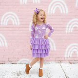 Child in a purple dress standing in front of a pink wall with white rainbows.