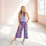 Woman in a purple top and floral pants standing in a sunlit room with high ceilings.