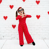 Person in a red outfit with heart patterns against a white brick wall with red hearts.