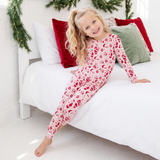 A young girl with blonde hair in a ponytail sits on a white bed wearing a festive red and white Santa Claus patterned long-sleeve pajama set. The room is decorated for Christmas with green garland draped over the headboard and red and green pillows.